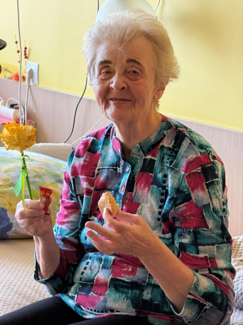 Iedzīvotāja raibā blūzē, sirmiem matiem sēž savā gultā ar ziedu un cepumu rokās. Smaida.