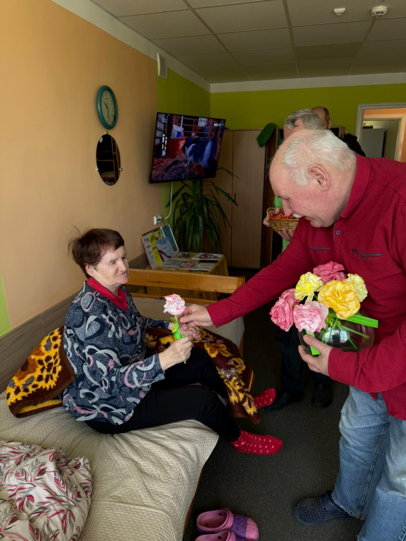 Smaidīgs kungs pasniedz ziedu iedzīvotājai, kura sēž savā gultā.