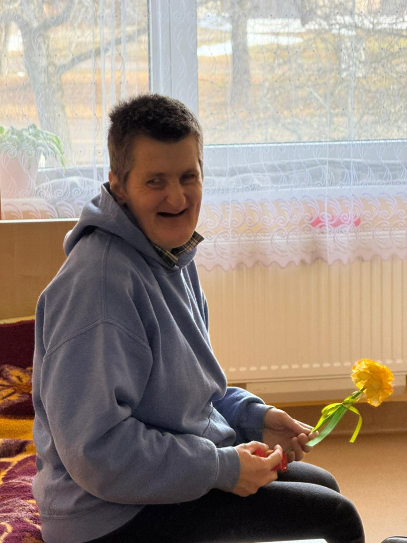 Iedzīvotāja sēž savā gultā ar dzeltenu ziedu rokās. Smaida.