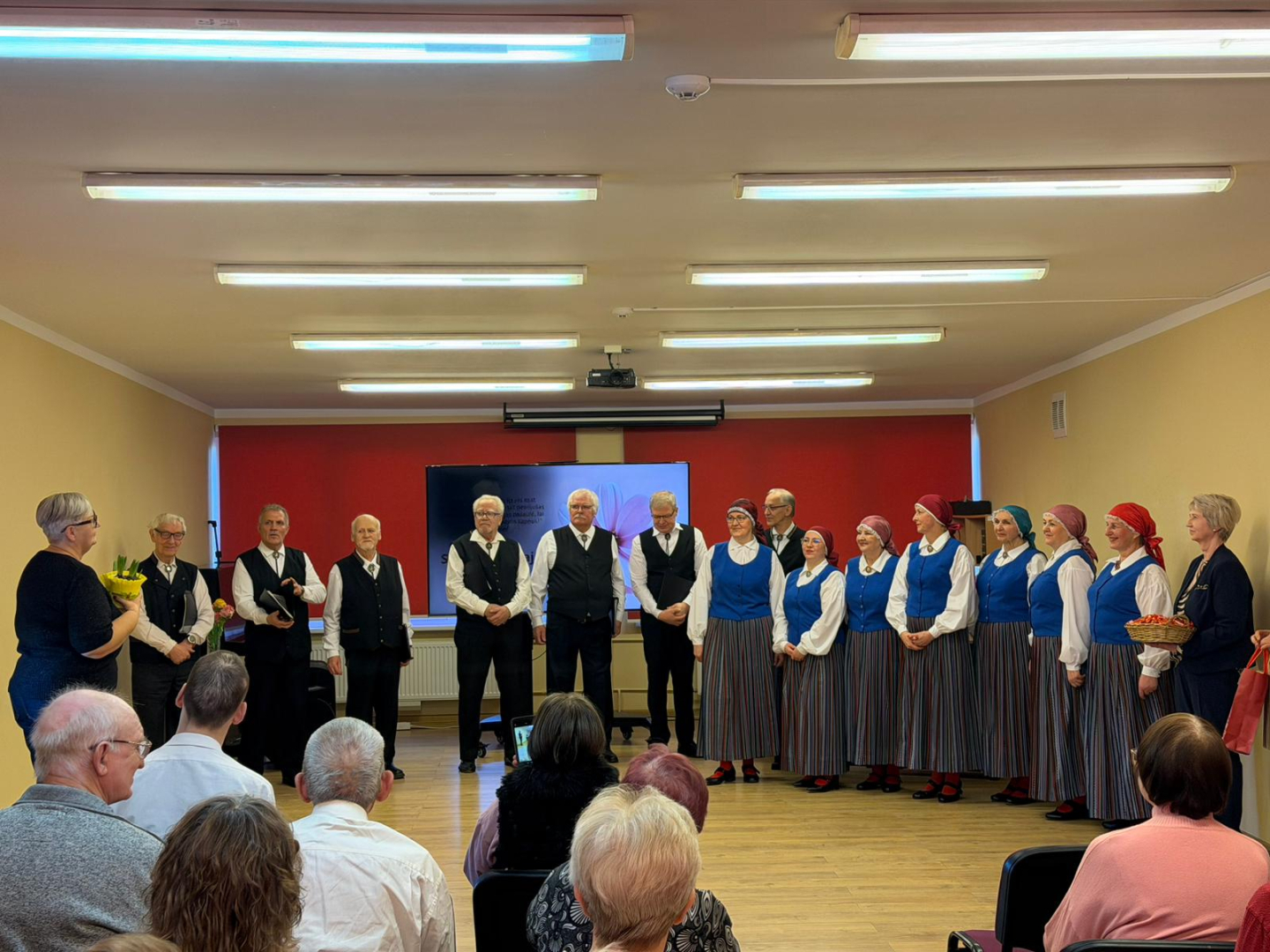 Iedzīvotāji sēž zālē, uz skatuves viesi, dāmu deju kolektīvs un kungu dziesmu ansamlis