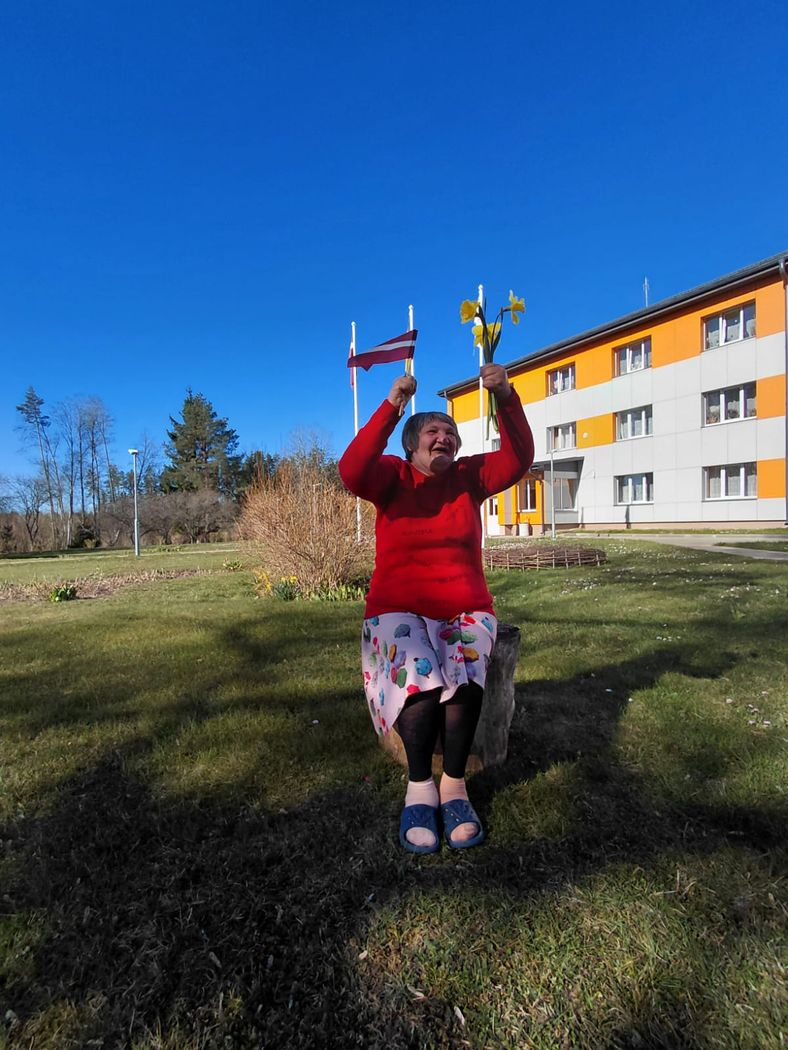 Kundze priecīga. Vienā rokā Latvijas karodziņš, otrā rokā narcises