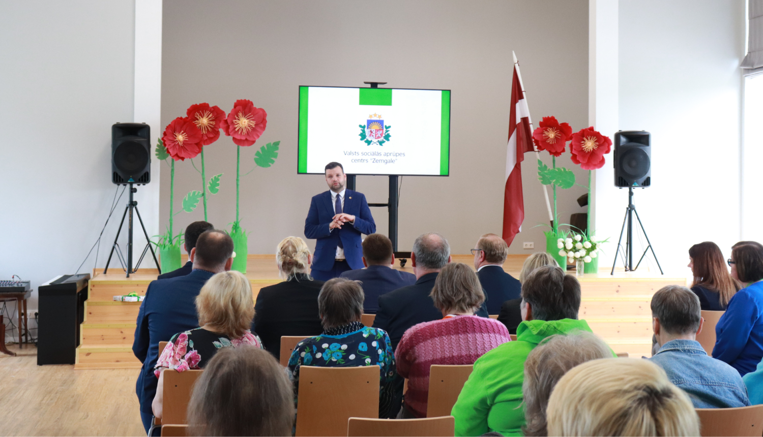 Labklājības ministrs stāv zāles priekšā un uzrunā klausītājus