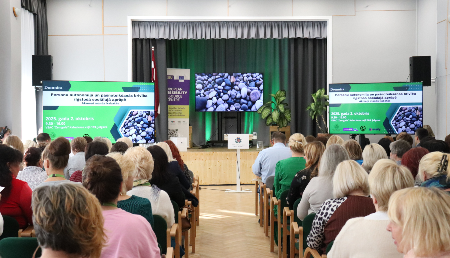 Skats no skatītāju zāles uz skatuvi ar ekrāniem un akmeņu attēliem