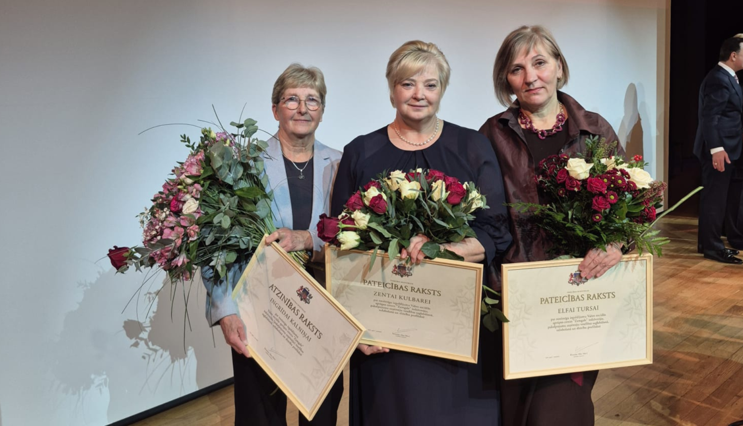 Fotogrāfijā trīs iestādes darbinieces ar ziediem un apbalvojumiem rokās