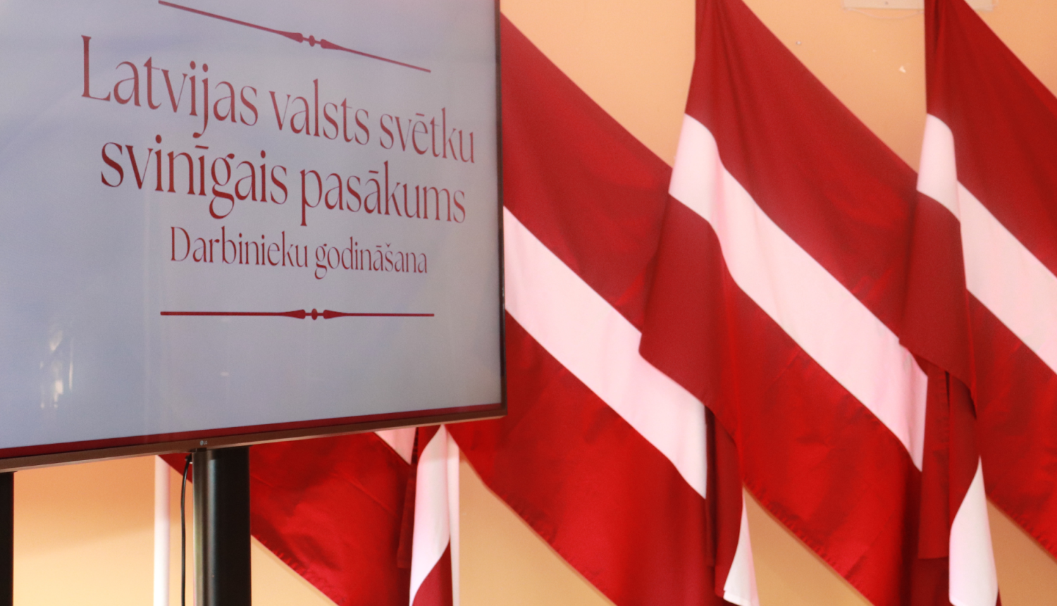 Attēlā ekrāns ar pasākuma baneri un fonā Latvijas karogu rinda