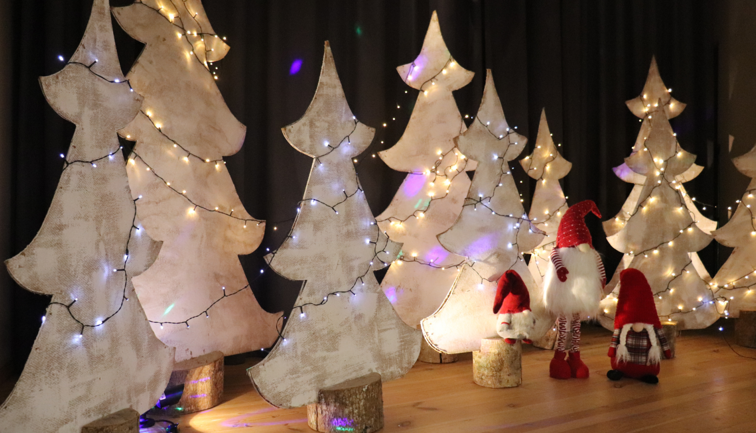 Attēlā skatuves dekorācijas. Trīs rūķi sarkanajos tērpos un rinda ar baltām eglēm, rotātā ar gaismiņām.