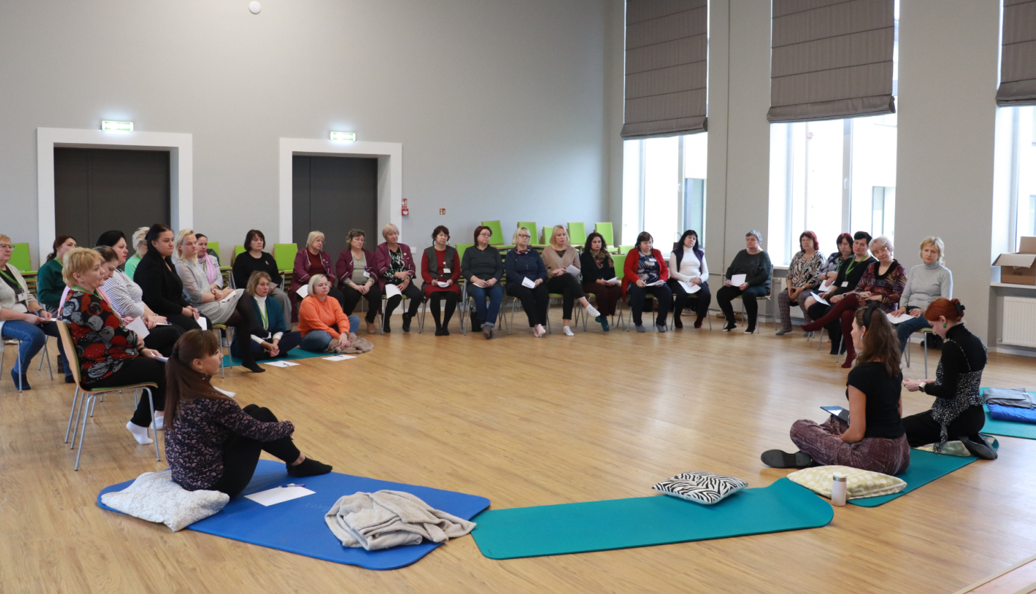 Pasākuma dalībnieki zālē sēž krēslos, kuri izkārtoti aplī, priekšā divas mākslas terapeites vada nodarbību