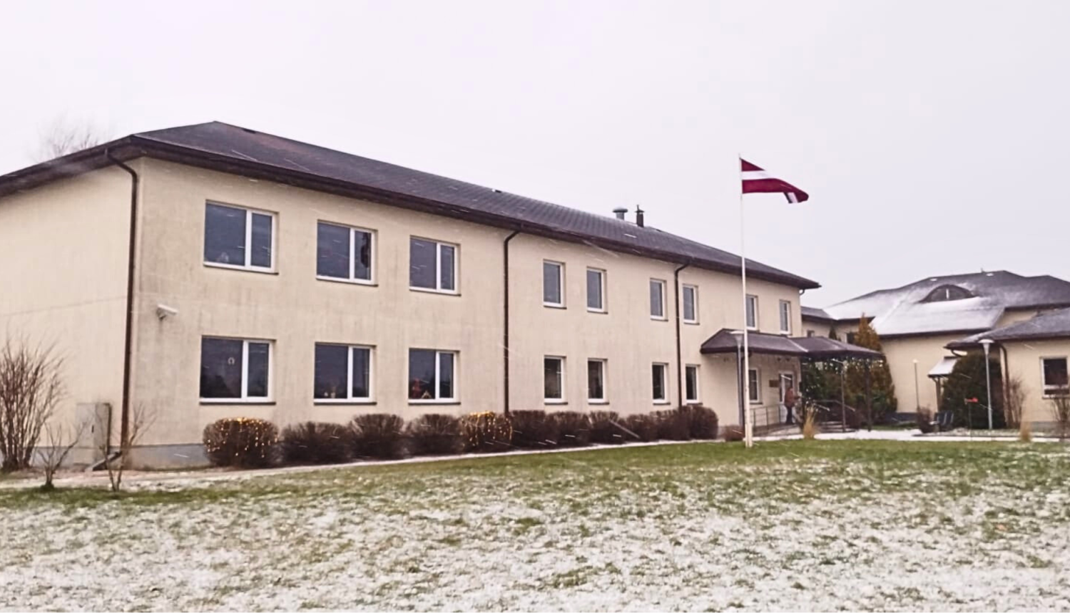 Grupu mājas divstāvu ēka bēšā krāsā, pie ieejas mastā plīvo valsts karogs