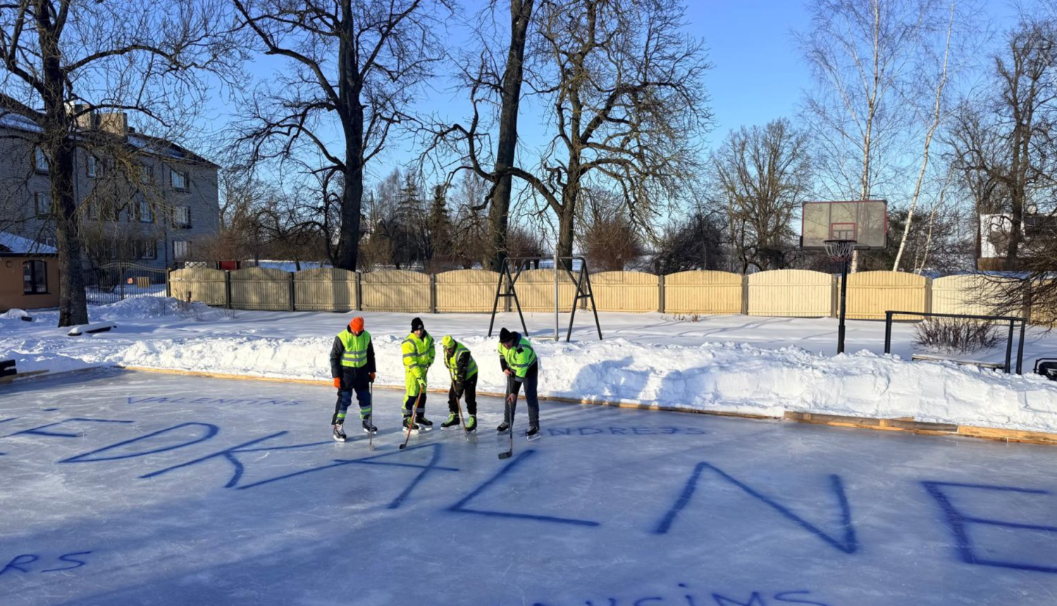 Četri hokejisti spilgti dzeltenajos tērpos stāv uz ledus ar nūjām. Uz ledus liels uzraksts ZIEDKALNE ar zilu krāsu