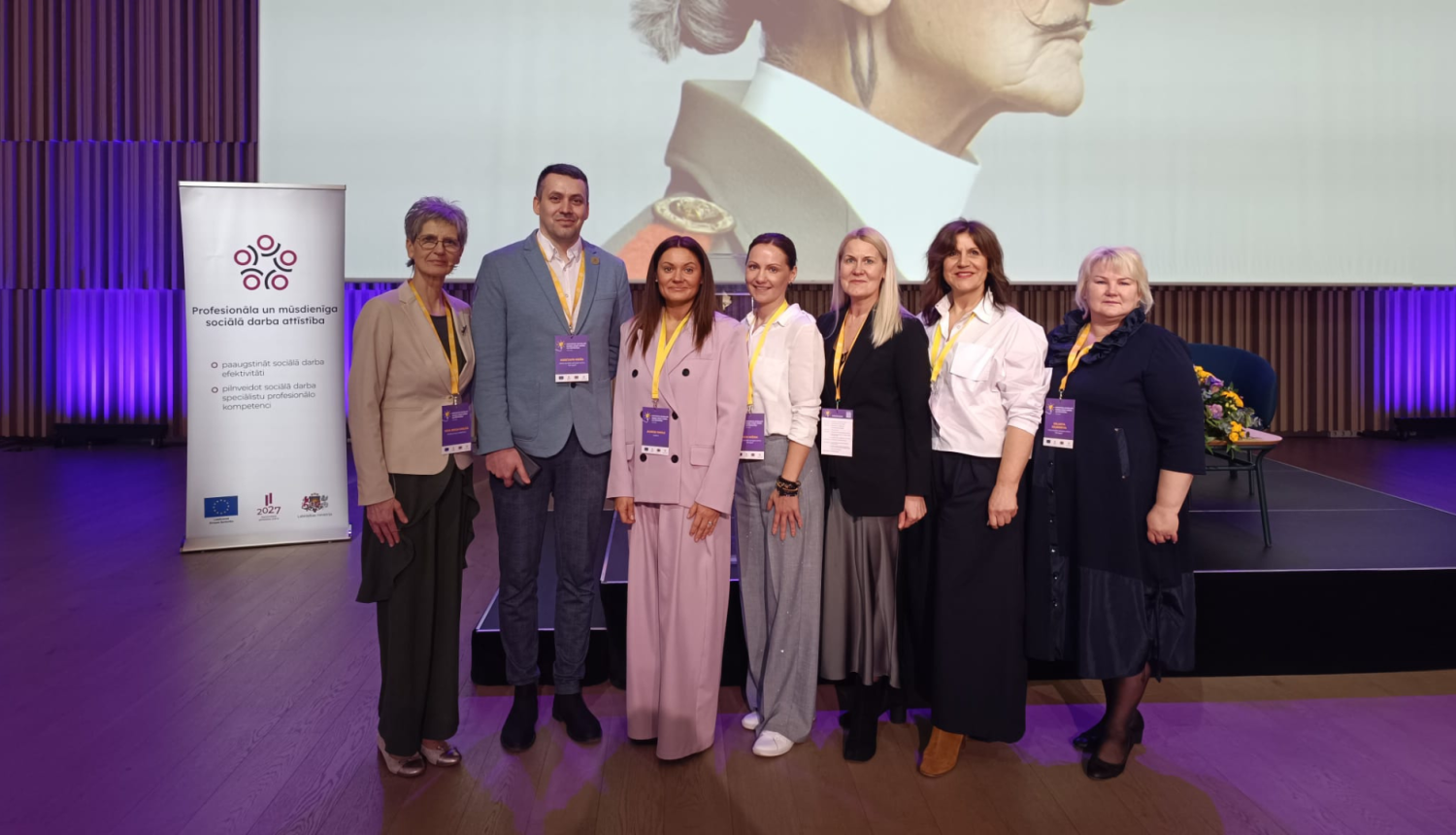 Iestādes darbinieku kopbilde. Septiņi cilvēki lietišķi ģērbušies stāv pie pasākuma skatuves.