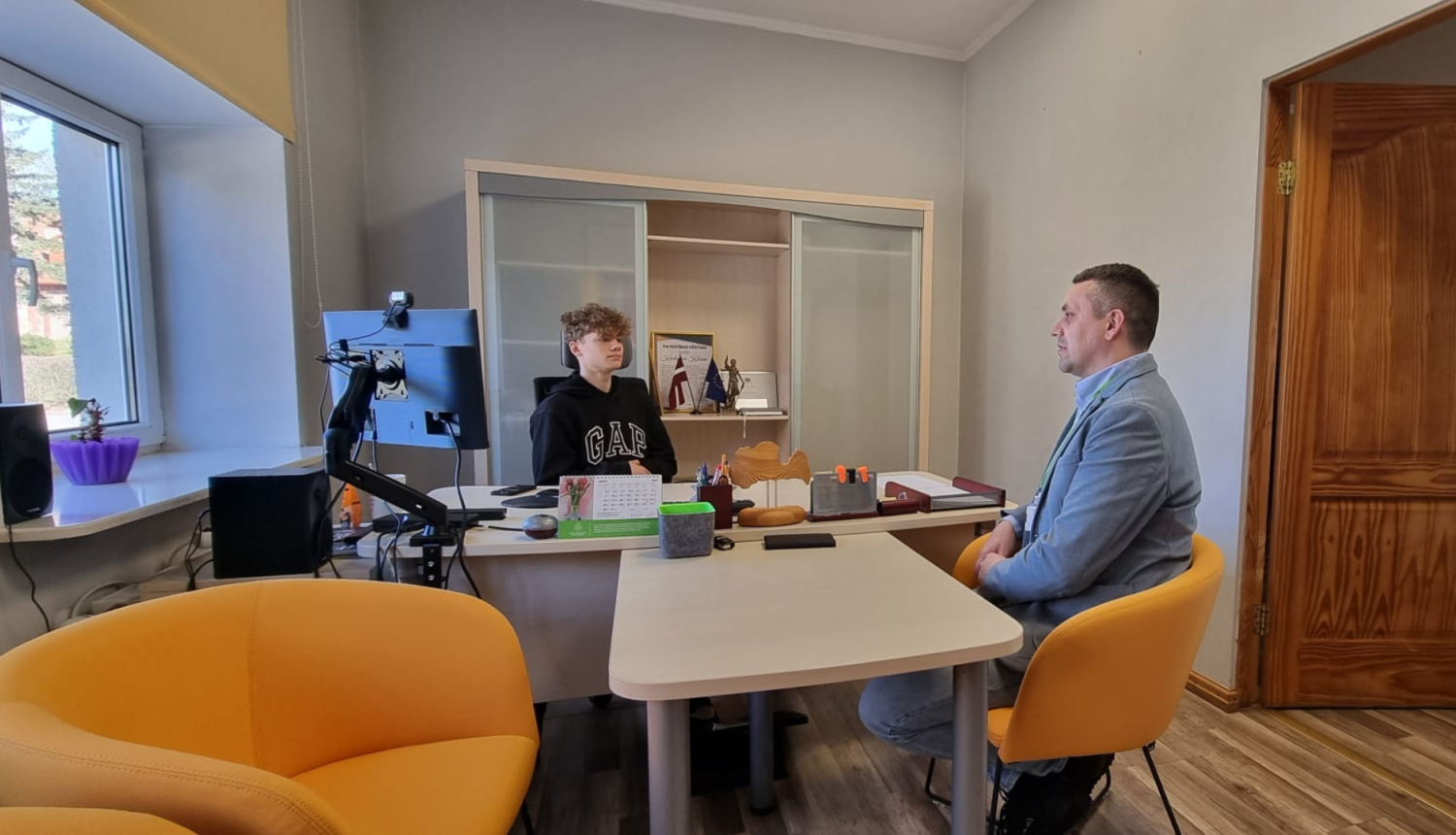 Kabinetā pie galda sēž iestādes direktors un jaunietis. Abi sarunājas.