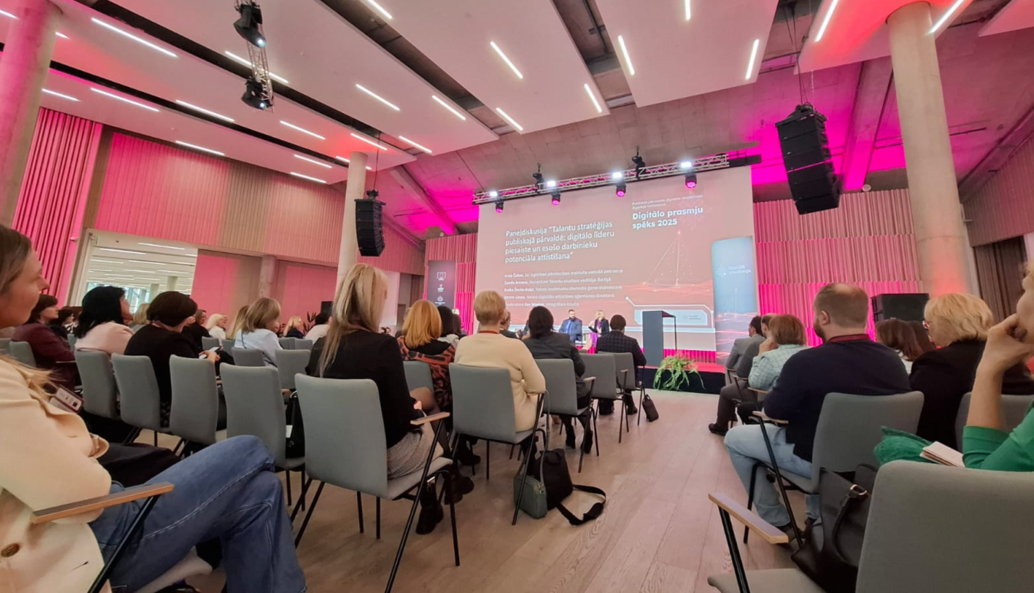 Fotogrāfija uzņemta Mežaparka Kokaru zālē. Attēlā konference, skats no skatītāju zāles, redzama pilna zāle ar konferences dalībniekiem, liels ekrāns un runātāji uz skatuves.