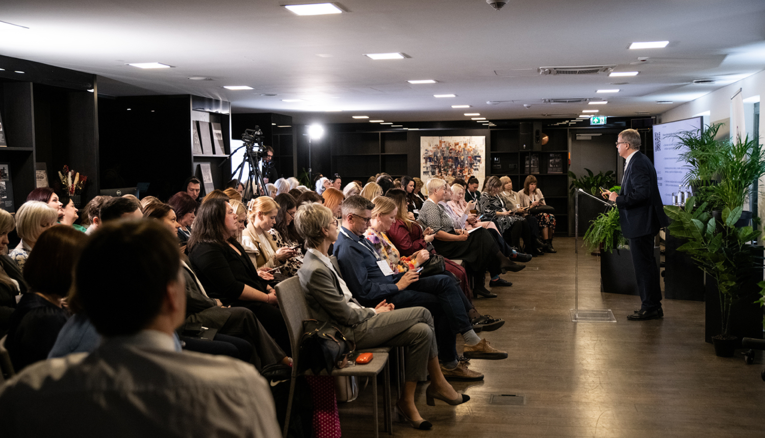 Konferences zāle pilna ar skatītājiem, kuri klausās vieslektora runā