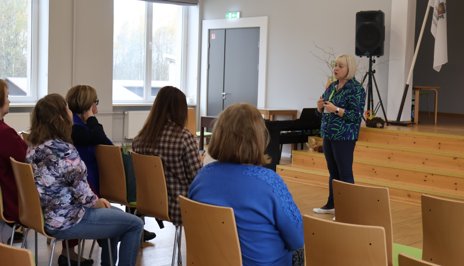 Zākē klausītāji sēž krēslos un klausās priekšā stāvošās mākslas terapeites stāstījumā