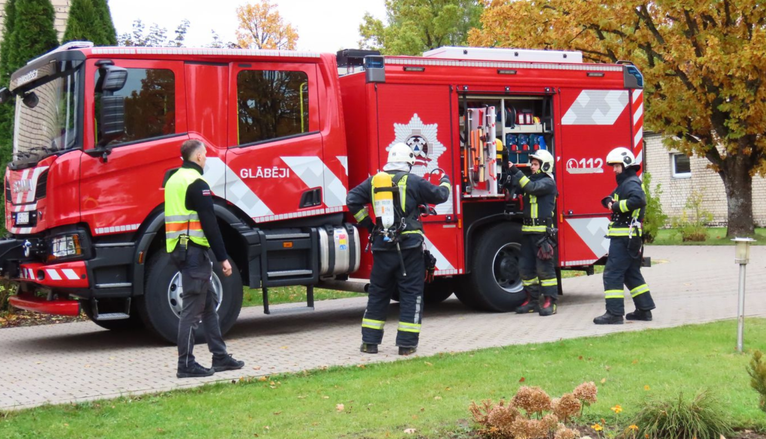 Attēlā četri ugunsdzēsēji pie ugunsdzēsēju mašīnas