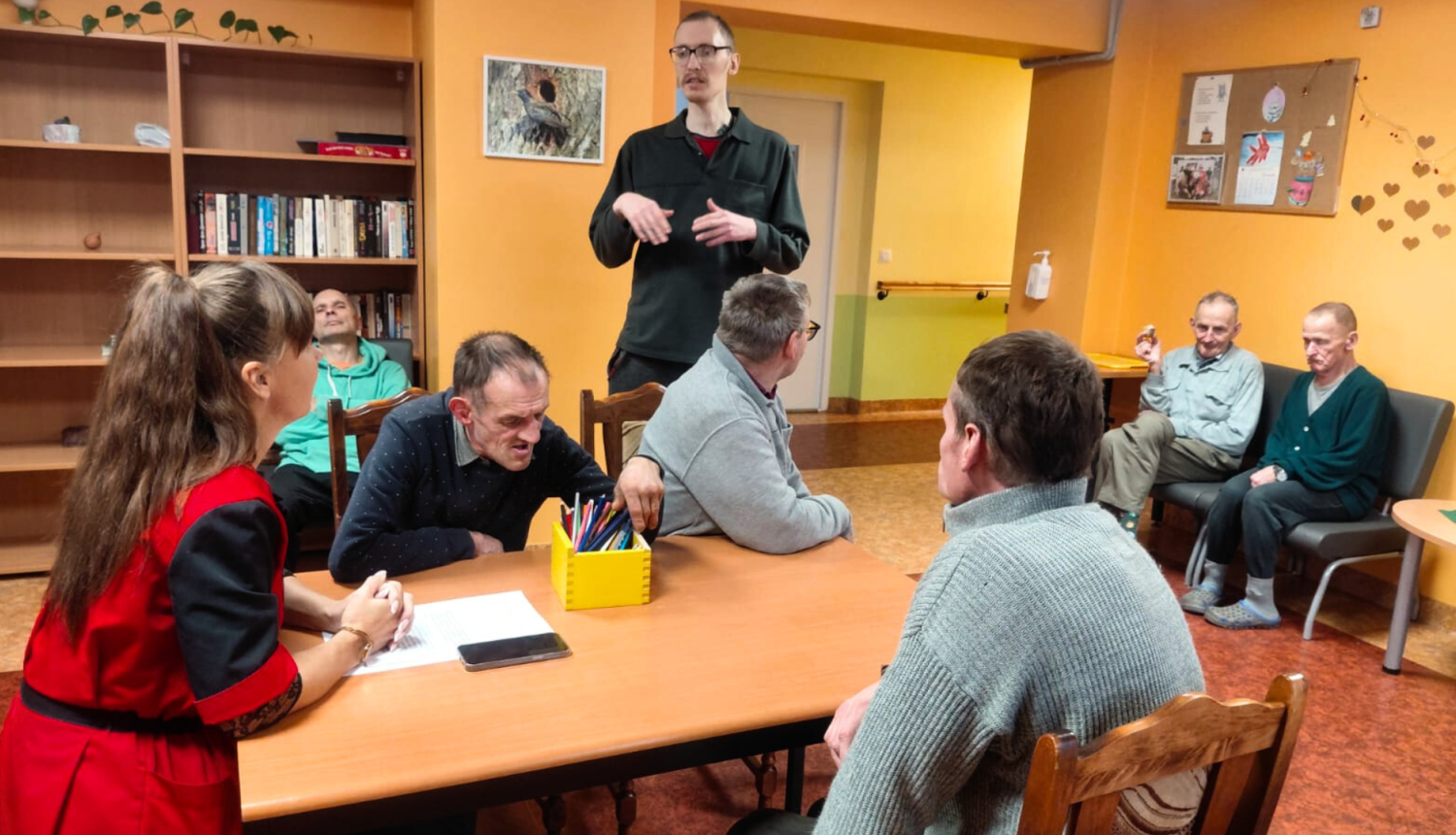 Attēlā darbiniece sarunājas ar iedzīvotājiem sēžot pie galda